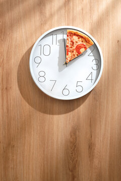White wall clock with a slice of pizza instead of hands on a light wooden table in soft sunlight. Funny creative Pizza Time concept.
