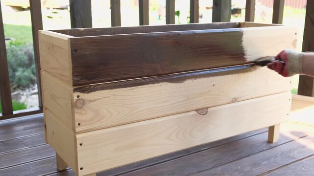 Brown oil for protection of wood by staining. Painting fence for protecting and pests. Woodworker applying finish to pine board box. Repairman varnishing with brush wooden fencing wall plank surface.