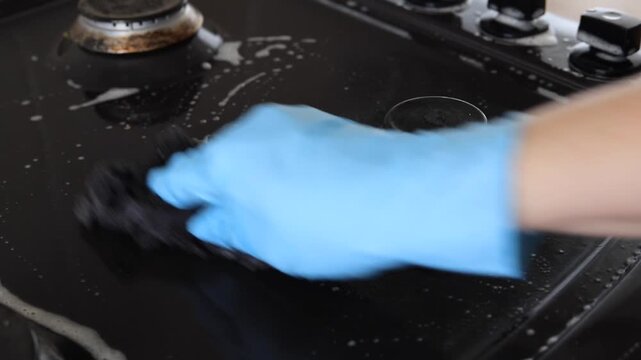 Deep cleaning gas stove burner from heavy grease, hand in rubber glove scrubbing black metal cooktop with soapy sponge