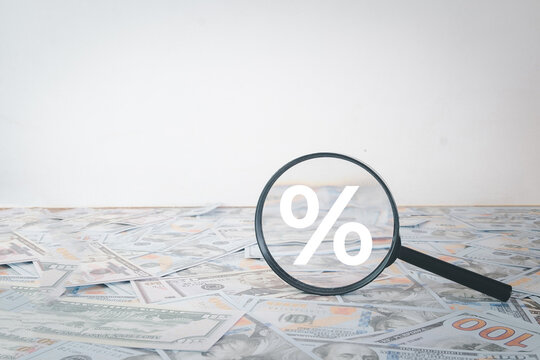 Interest rate and financial analysis concept. Magnifying glass with a percentage sign resting on a pile of US dollar bills against a white background with copy space for text.