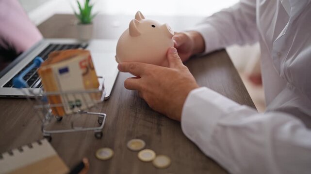 Man in a white shirt sitting at an office desk putting euro coins into a piggy bank, symbolizing financial planning, investment strategies, and saving money for future retirement security