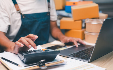Person wearing an apron uses a calculator and laptop while managing paperwork and packages, likely...