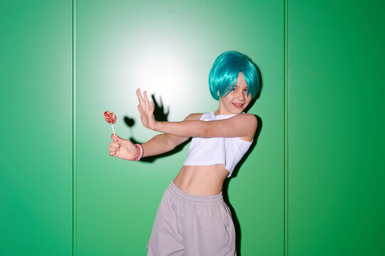 Young person with vibrant blue wig and lollipop strikes a playful pose against a bright green wall