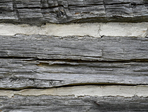 Horizontal close-up view of a weathered log cabin wall with flat logs that show dried wood with visible splintering and cracking and with porous, uneven chinking between the logs