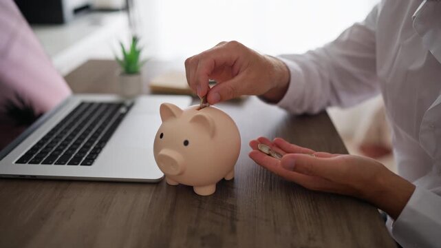 Unrecognizable businessman sitting at his office desk inserting coins into piggy bank, symbolizing financial savings, investment growth, retirement planning, and budget management