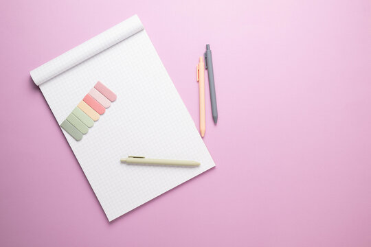 A note pad with blank grid paper on a bright purple background with multicolored sticky note tabs and pastel pens.