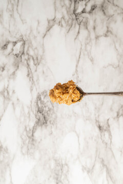 Silver, antique spoon full of crunchy, creamy peanut butter, above a gray and white marbled surface, vertical
