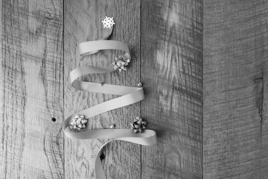 Black and white image, a ribbon Christmas tree shape with bows and ornaments on a wooden background with copy space.