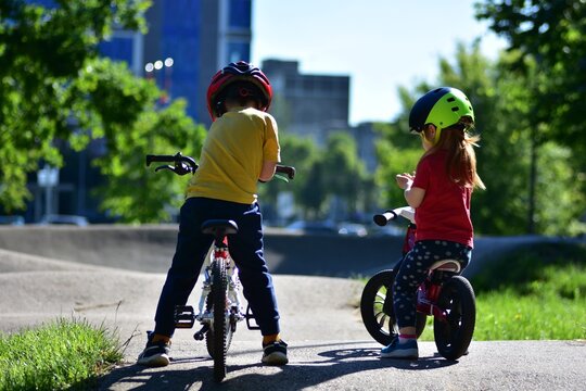 Two young children wearing protective helmets pause with their bicycles on a pump track in a sunny urban park. Active childhood, safe cycling, friendship, balance training and healthy recreation