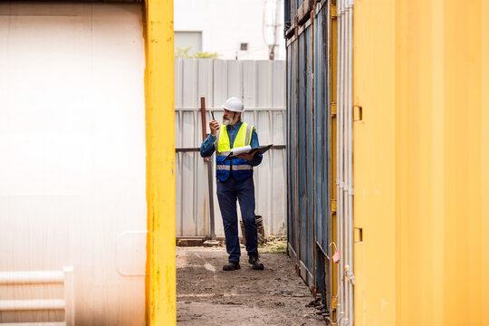 Safety inspector in hard hat and high-visibility vest checks a container area, speaking on a radio and noting details on a clipboard between yellow cargo units.