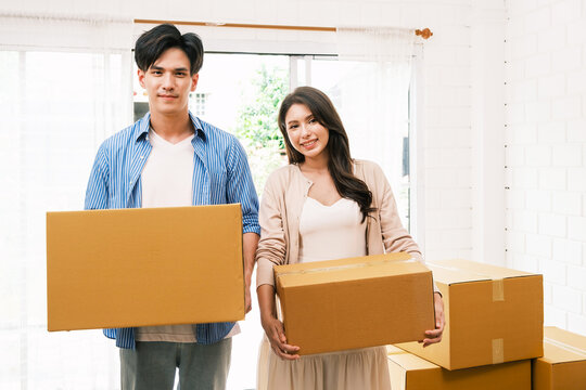 Two adults indoors holding cardboard boxes amid stacks, suggesting a move into a bright new home.