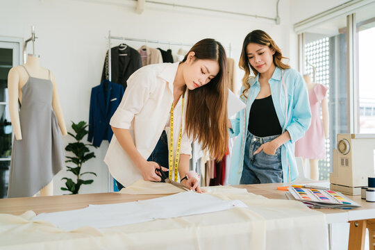 Two fashion designers work in a bright studio; one cuts fabric on a pattern while the other checks colors, surrounded by mannequins, tools, and a sewing machine.