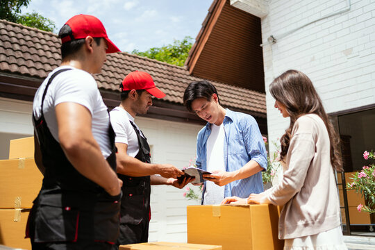 Moving crew and clients outside a house review a checklist and sign delivery paperwork beside stacked cardboard boxes.