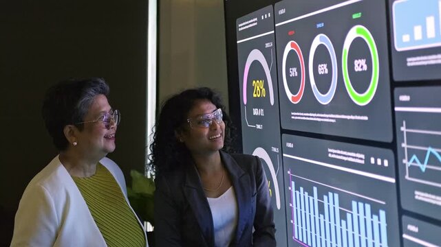 Two professionals review a large digital dashboard with charts and metrics, discussing performance analytics and insights in an office setting.
