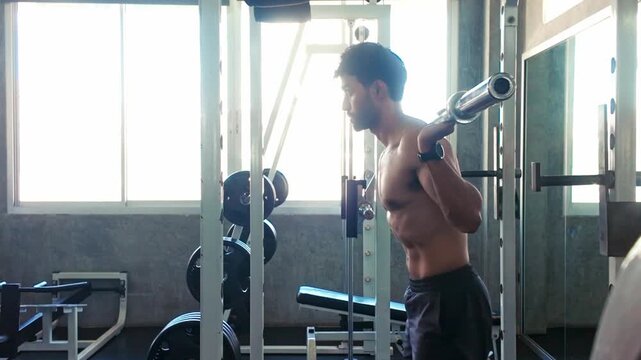 A man performs a barbell back squat in a gym, leaning forward under the bar inside a squat rack with weights and mirrors, lit by window light.