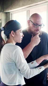 Two colleagues in a bright studio review something on a tablet; the woman gestures while the man listens thoughtfully, suggesting coaching, planning, or a review session.
