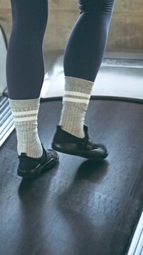 Close-up of a person walking on a treadmill, wearing dark leggings, white athletic socks, and black athletic shoes in a gym.