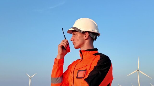 Engineer in orange high-visibility jacket and hard hat uses a walkie-talkie at a wind farm under a clear blue sky