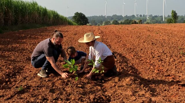 Two adults and a child plant a young tree in a plowed field as wind turbines spin on the horizon, highlighting sustainable agriculture and renewable energy.