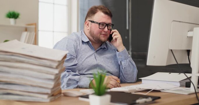 Man at office desk on phone call. Wearing glasses, he manages a stack of paperwork and documents beside a computer, taking notes working to meet deadlines. Clear busy office work theme.