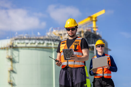 Engineer using tablet and walkie talkie at LNG plant facility, Technician team checking data on device at natural gas terminal, Worker in safety gear managing operations at industrial storage site