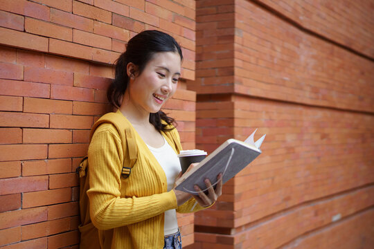 Young asian female student with backpack smiling while holding a book and a takeaway coffee cup, walking outside a modern building university campus