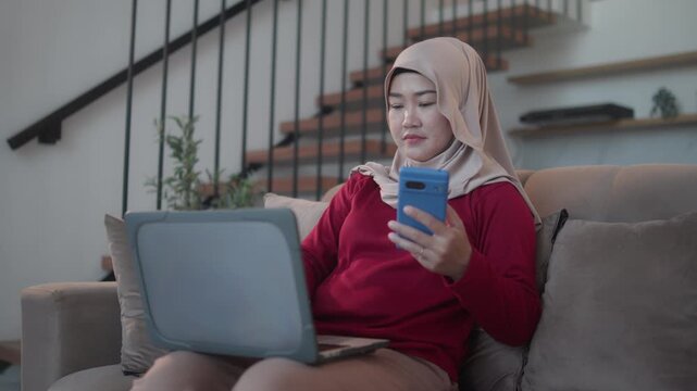 A woman wearing a hijab sits on a living room sofa while using a laptop and smartphone for two-factor authentication, illustrating online security, secure login, and digital banking.