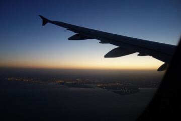 Fototapeta premium View of an airplane wing from a passenger window during flight above the sea at twilight. Aerial travel scene featuring a coastline with illuminated city lights, a clear gradient sky, and the horizon 