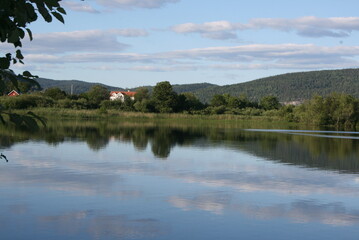 Peaceful lake with reflections of forest and clouds, a small countryside house and calm summer landscape. Natural outdoor scenery with copy space for travel and nature themes. © GlaDDeN