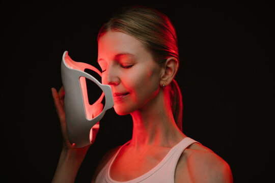 infrared light therapy mask. woman is holding the mask with closed eyes her face is illuminated by red light. black  background. Contrast red light. Horizontal composition