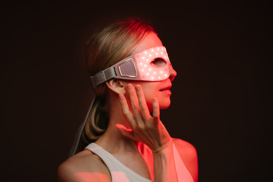 Red light therapy eye LED mask. woman profile wearing infrared light therapy mask. touching the mask with her hand.  looking side. dark background