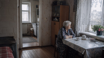 Fototapeta premium Elderly woman sitting alone at a kitchen table by the window in a modest rural home