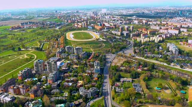Aerial view of the La Maura trotting racecourse in the San Siro district of Milan, Italy, with the sand track, training rings, residential towers, parks and the city skyline in spring.