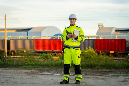Professional logistics engineer in high visibility safety uniform using laptop and radio at container yard, Industrial worker managing freight transport operations at container terminal