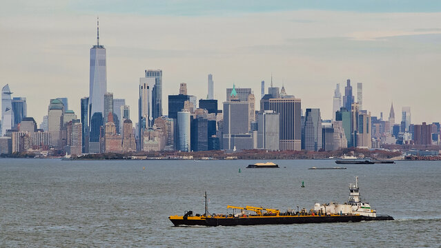 Panoramic view of Lower Manhattan skyline with Freedom Tower skyscrapers and an industrial tugboat barge vessel sailing across New York Bay on an overcast day