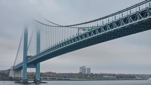 Low angle view of a massive steel suspension bridge disappearing into thick fog and mist on an overcast cloudy day over Verrazzano Narrows in New York City