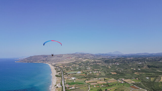 Aerial view of a paramotor paraglider flying high over the Crete island coastline with blue sea and beach landscape in Greece on a clear sunny summer day