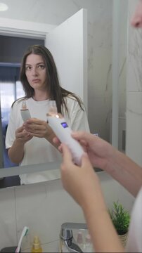 A young woman stands in front of the bathroom mirror during her morning skincare routine, holding a facial pore vacuum cleanser and selecting the preferred cleaning mode before beginning her daily bea