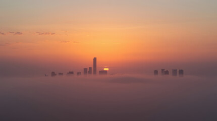 Fototapeta premium Misty sunrise skyline with distant skyscraper peeking above thin morning fog, serene light