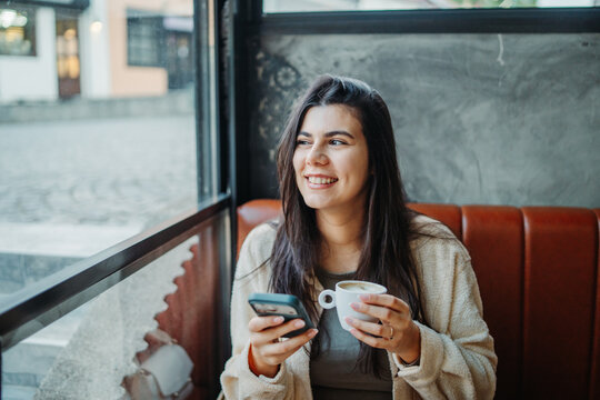 Woman enjoying coffee with smartphone in cafe looking out window