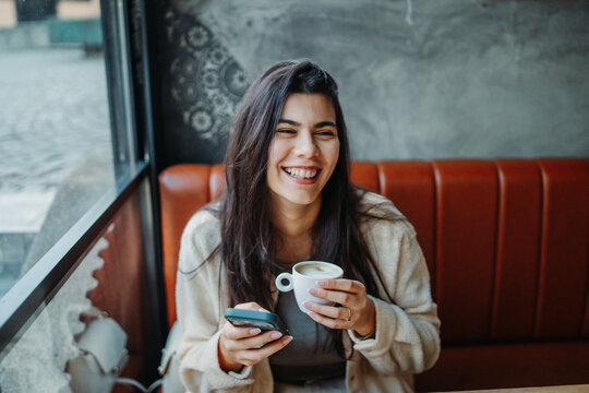 Woman smiling, drinking coffee, and using smartphone in cafe