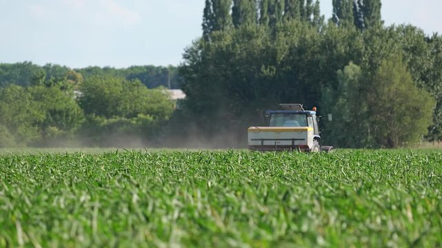 Agricultural tractor operating in green crop field during summer with rural landscape and drifting dust in the background.
