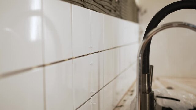 Close-up view of a construction worker's hands placing white ceramic tiles on a wall coated with adhesive, meticulously inserting plastic spacers for perfect alignment during a kitchen remodel