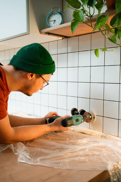 Man wearing a green beanie and glasses diligently drilling a circular hole into white ceramic kitchen wall tiles with a power tool, undertaking a home renovation project