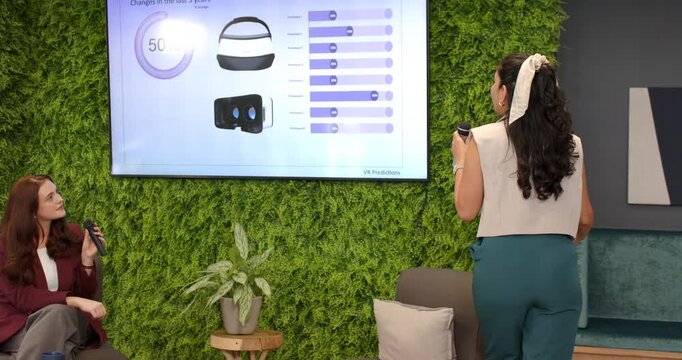 Two women in office host holding mic pointing to slide charts explaining headset data, copy space