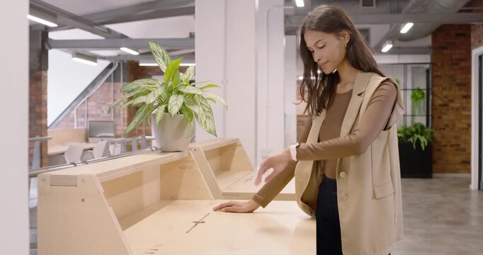 Woman in beige vest walking to loft counter after seeing plant, pausing, checking watch, smiling