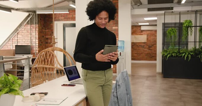 African American man leaning at desk with laptop, scrolling tablet, finishing review and tucking it