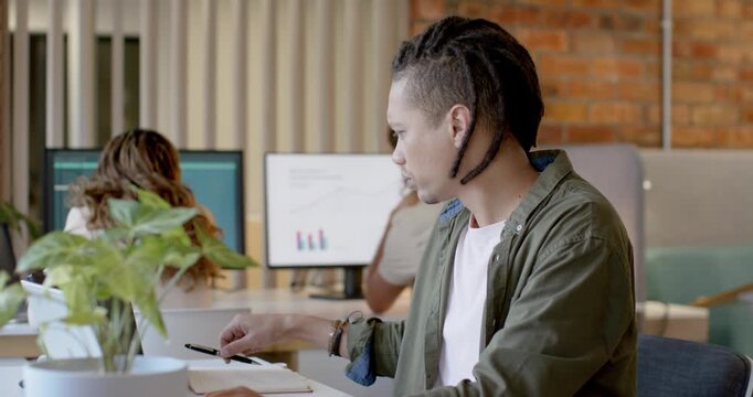 Asian male working on laptop at desk, glancing down, reaching for phone and writing in notebook