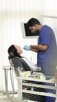 A male dentist in blue scrubs examines a female patient's open mouth with dental instruments in a modern clinic