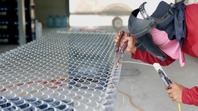 Welder wearing a protective face shield and gloves welds a steel rebar grid on the ground, with sparks and workshop tools visible in the background. Represents construction, metalwork, and industrial
